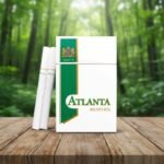 ซองบุหรี่ Atlanta Menthol สีขาวแถบเขียว วางคู่กับมวนบุหรี่บนโต๊ะไม้ ท่ามกลางบรรยากาศสวนสีเขียว