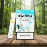 ซองบุหรี่ Raison French Yogo ลายหอไอเฟลสีฟ้าขาว พร้อมมวนบุหรี่ วางบนโต๊ะไม้ในบรรยากาศป่าธรรมชาติ
