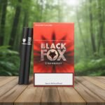 ซองบุหรี่ Black Fox Strawberry วางคู่กับมวนบุหรี่สีดำบนโต๊ะไม้ ฉากหลังเป็นป่าธรรมชาติ