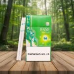 ซองบุหรี่ ESSE Menthol Plus+ สีเขียวสดใสวางบนโต๊ะไม้ พร้อมมวนบุหรี่สลิมโชว์เม็ดบีบ ฉากหลังเป็นป่าไม้