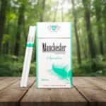 ซองบุหรี่ Manchester Menthol SuperSlim สีขาวคาดเขียวมิ้นต์ ดีไซน์ทันสมัย วางบนโต๊ะไม้ฉากหลังป่าธรรมชาติ