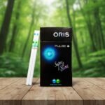 ซองบุหรี่ Oris Pulse Fusion Mint สีดำดีไซน์ทันสมัยพร้อมกราฟิกวงกลมสีฟ้าและเขียว วางบนโต๊ะไม้ฉากหลังป่า