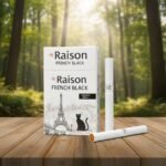 บุหรี่ Raison French Black ซองสีขาวลายกราฟิกแมวดำและหอไอเฟล วางบนโต๊ะไม้ฉากหลังป่าไม้ธรรมชาติ