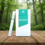 บุหรี่ Atlanta Green Super Slim ซองสีขาวคาดแถบสีเขียวพาสเทล วางคู่กับมวนบุหรี่ซุปเปอร์สลิมบนโต๊ะไม้ฉากหลังป่าไม้