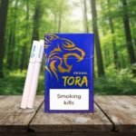 บุหรี่ Tora Original ซองสีน้ำเงินลายเสือสีทอง มวนบุหรี่สีขาว วางบนพื้นไม้ฉากหลังป่าสีเขียว