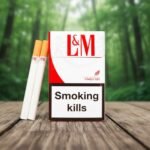 ซองบุหรี่ L&M Red Fine Cut สีขาวแดงเกรดพรีเมียมจากสวิตเซอร์แลนด์ วางบนโต๊ะไม้ในบรรยากาศธรรมชาติ พร้อมมวนบุหรี่ก้นกรองสีส้มคลาสสิก