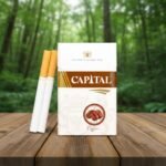 ซองบุหรี่ Capital Coffee สีขาวคาดน้ำตาลทอง พร้อมรูปเมล็ดกาแฟ วางบนโต๊ะไม้ในบรรยากาศธรรมชาติ