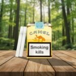 บุหรี่ Camel Non-Filter แบบไม่มีก้นกรอง ซองสีเหลืองคลาสสิก วางบนโต๊ะไม้ฉากหลังป่าเขียวขจี