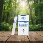 บุหรี่ Manchester Queen Aqua ซองสีขาวแถบน้ำเงิน มวนขนาดควีนไซส์ วางบนโต๊ะไม้ฉากหลังป่าเขียวขจี