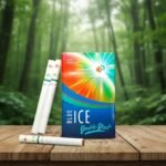 บุหรี่ Blue Ice Double Blast Cool Summer ซองหลากสีลายแสงสีส้มเขียวและน้ำเงิน พร้อมมวนบุหรี่แบบ 2 เม็ดบีบ วางบนโต๊ะไม้ฉากหลังป่าธรรมชาติ