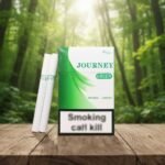 บุหรี่ Journey Green ซองสีขาวตัดเขียวลายกราฟิกใบไม้ มวนสีขาวสะอาดก้นกรองสีขาวแถบเขียว วางบนโต๊ะไม้ฉากหลังป่าธรรมชาติ