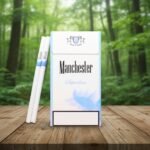 ซองบุหรี่ Manchester Blue SuperSlim สีขาวแถบฟ้า วางบนโต๊ะไม้ท่ามกลางบรรยากาศป่าธรรมชาติ