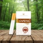 บุหรี่ Capital Coffee ซองสีขาวแถบน้ำตาลลายเมล็ดกาแฟ วางบนโต๊ะไม้ฉากหลังป่าสีเขียว