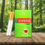 บุหรี่ Capital Double Apple ซองสีเขียวสดใสรูปแอปเปิ้ลคู่ พร้อมมวนบุหรี่วางบนโต๊ะไม้ในบรรยากาศป่า